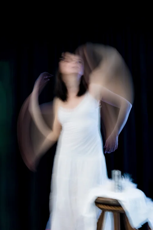 Femme en nuisette blanche sur fond noir qui bouge les bras, ce qui laisse des trainées lumineuses.