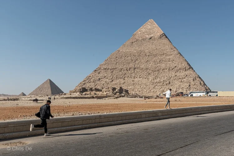 Deux hommes courent sur une route devant les pyramides alignées de Khephren, de Kheops et de Mykérinos.